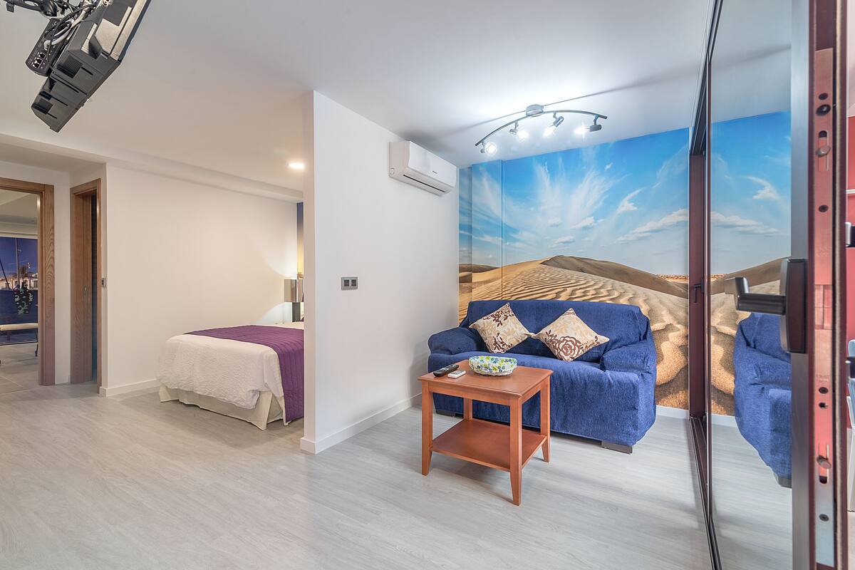 Une chambre d’hôtel moderne avec un lit, un canapé bleu, une petite table en bois et une fresque murale sur le thème du désert.