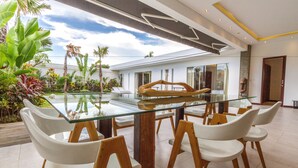 A modern dining area with a glass table and white chairs, surrounded by lush tropical plants. - Villa Saha (Seminyak)