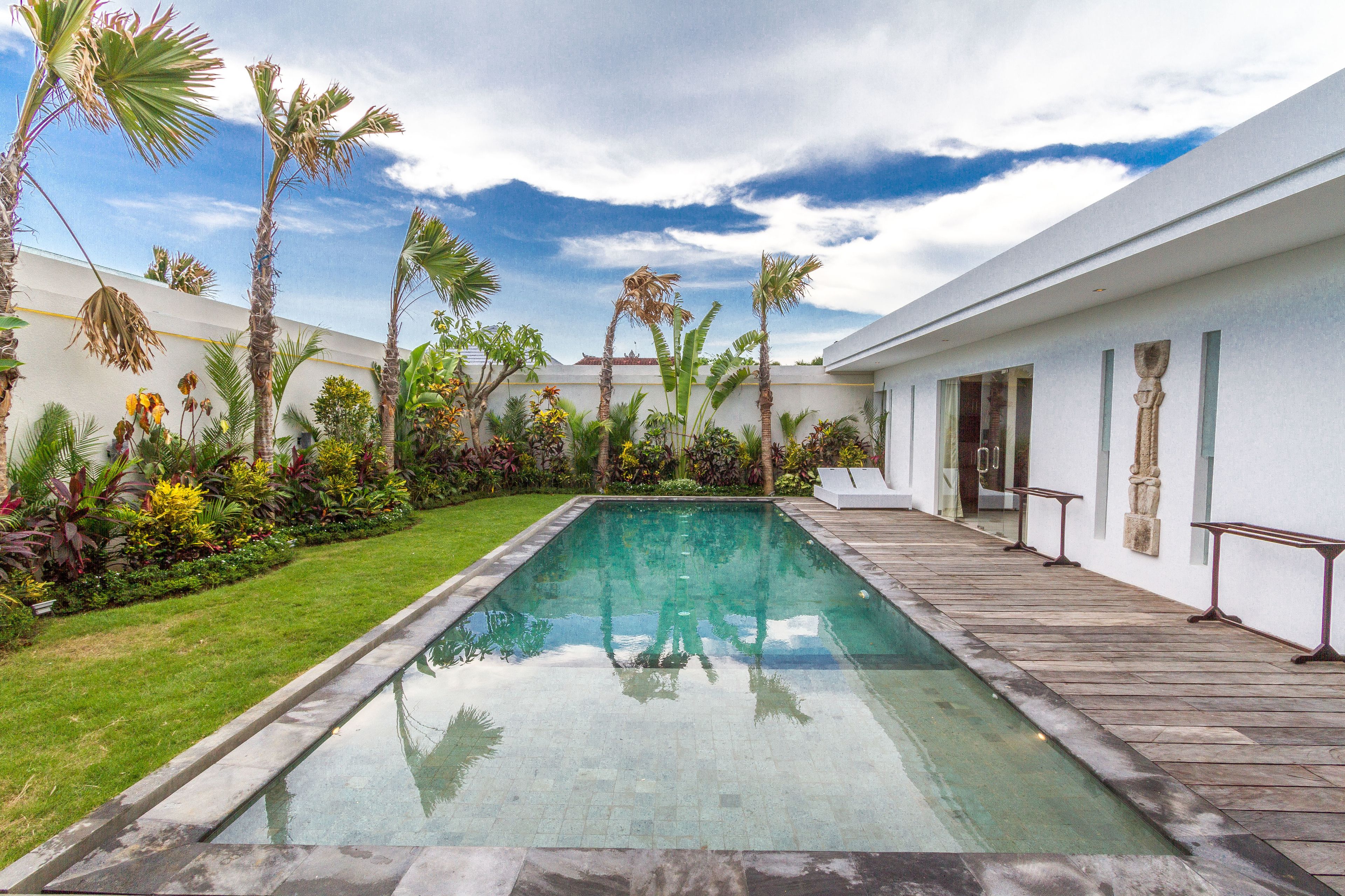 A rectangular swimming pool with a tiled deck, surrounded by palm trees and various plants, with a white building in the background.
