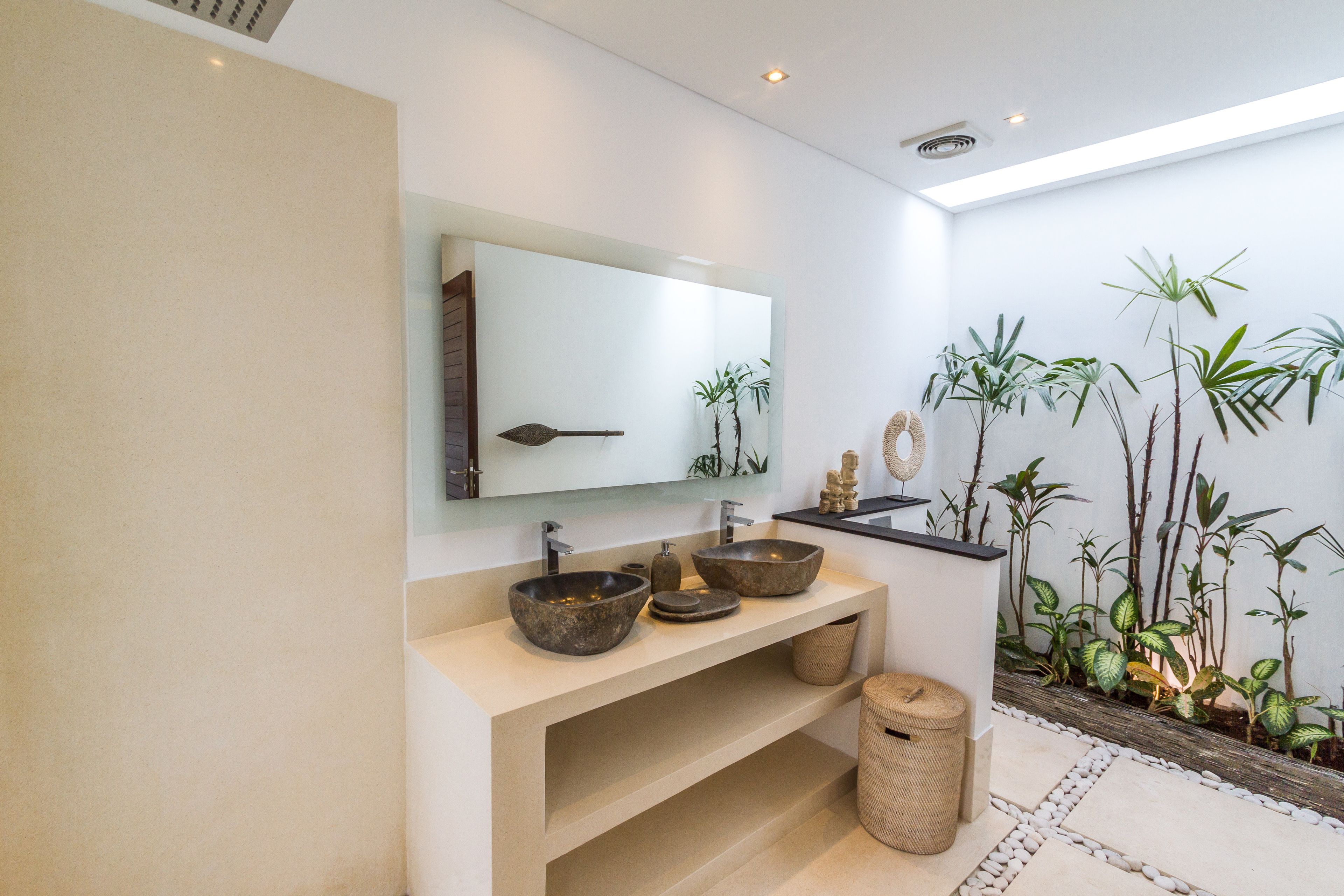 Un baño moderno con un espejo grande, dos lavamanos tipo recipiente y un pequeño estante. Hay plantas y un revestimiento decorativo en una pared.