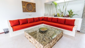 A modern living room with a red sectional sofa, a glass coffee table with a natural wood base, and a large mirror reflecting the room. - Villa Saha (Seminyak)