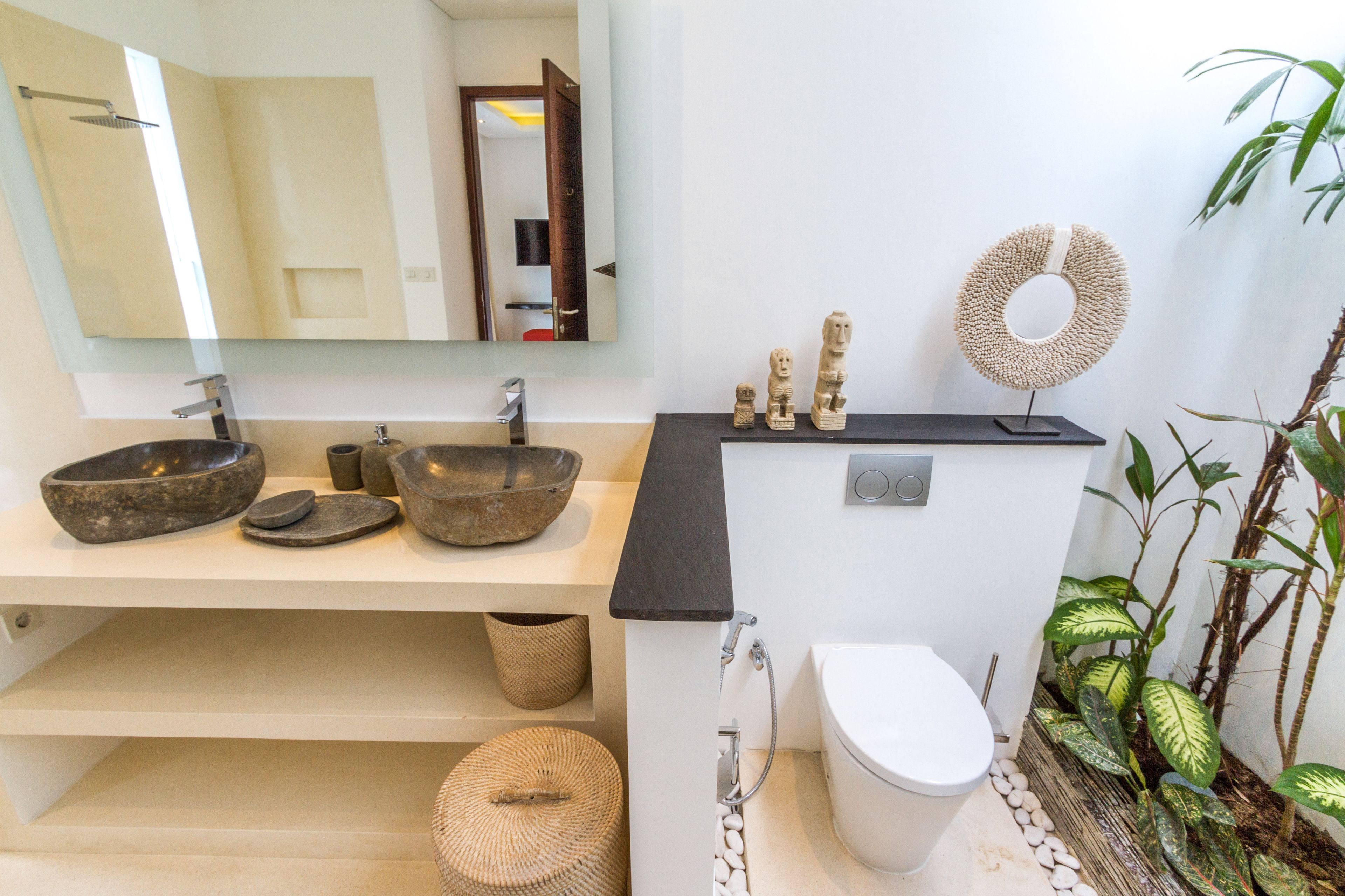 Un baño moderno con un espejo grande, dos lavamanos de piedra, una encimera negra, un inodoro y elementos decorativos como una cesta tejida y un adorno circular en la pared.
