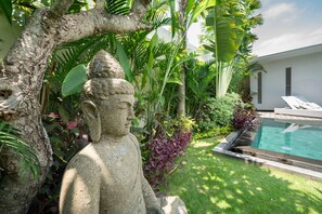 A statue in a lush garden with a pool and lounge chairs. - Villa Tira (Seminyak)