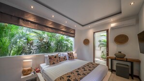 A modern bedroom with a large bed, a bedside table with a lamp, a small fireplace, and a view of lush greenery through the window. - Villa Tira (Seminyak)