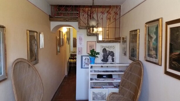 A hallway with framed artwork on the walls, a white shelf with books, and wicker chairs. - Affittacamere Grandpa Mau (Lucca)