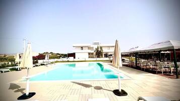 A swimming pool with lounge chairs and umbrellas, a covered outdoor seating area, and a building in the background.