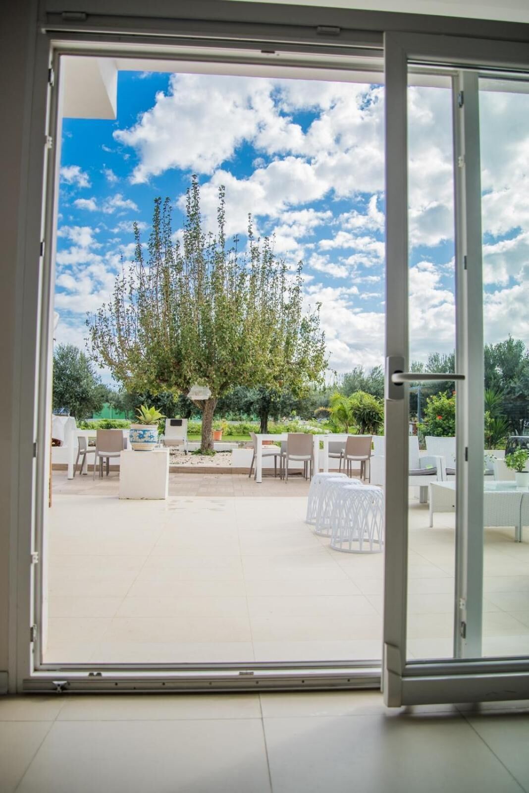 A view through a glass door into an outdoor patio area with a large tree, white outdoor furniture, and a clear blue sky with clouds.