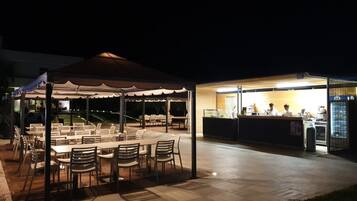 Un espace de restauration en plein air avec des tables et des chaises sous une marquise, un bar avec un comptoir et du personnel, et une terrasse bien éclairée.