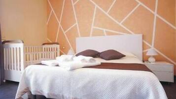 A neatly made bed with white linens and two dark pillows, a white bedside table with a lamp, and a radiator on the left.