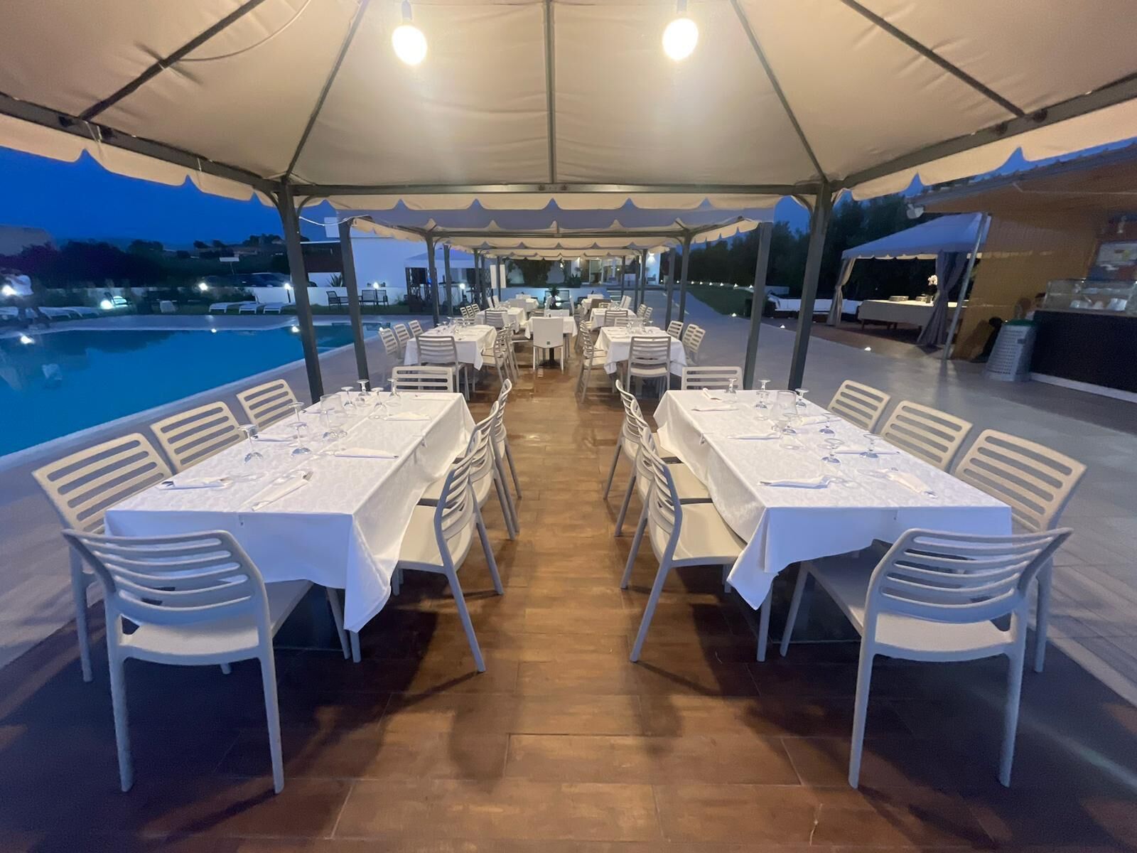 Un área de comedor al aire libre con mesas y sillas bajo un toldo, con vista a una piscina.
