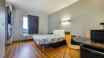 A hotel room with a bed, a desk, a chair, a flat-screen TV, a heater, and a window with curtains.