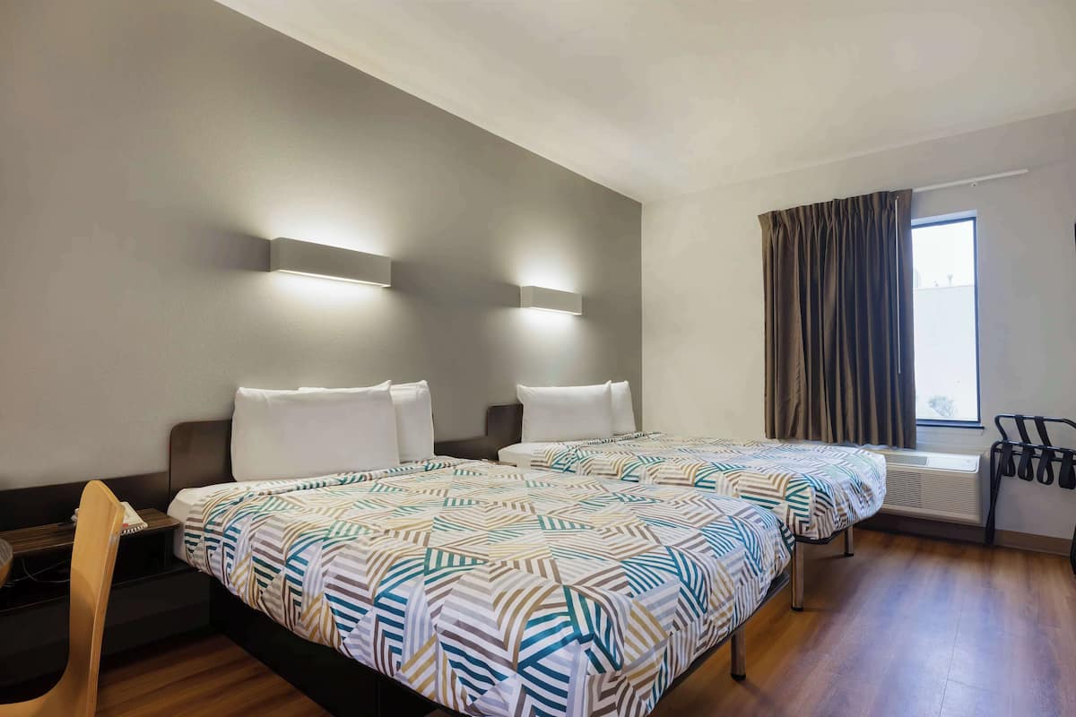 Een hotelkamer met twee bedden, een bureau, een stoel, een raam met gordijnen en een wandairconditioning.