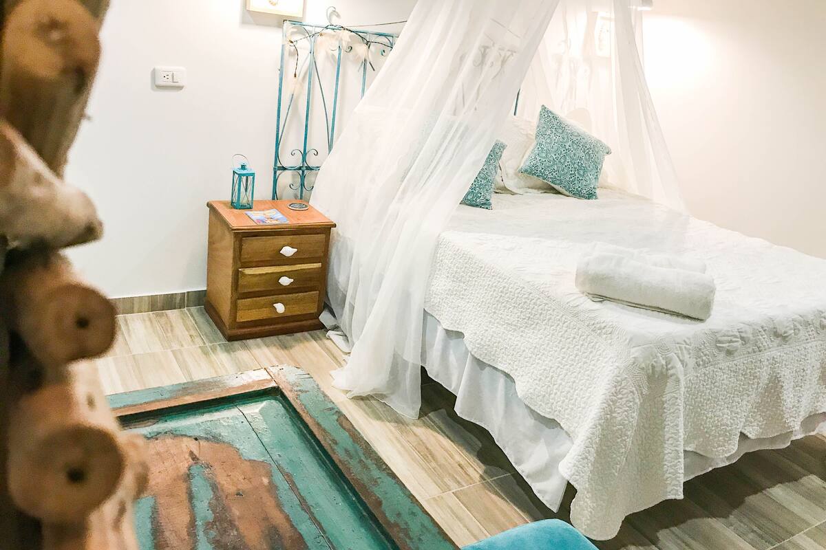 Une chambre à coucher avec un lit à baldaquin, une table de chevet en bois, une petite table sur laquelle est posée une lanterne, et une chaise bleue.