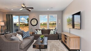 A living room with two sofas, a coffee table, a TV, and a large window with a view of a desert landscape. - Another Day In Paradise (St. George)