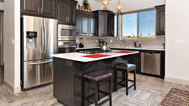 A modern kitchen with dark cabinets, stainless steel appliances, a large island with stools, and pendant lighting. - Red Rock Retreat (St. George)
