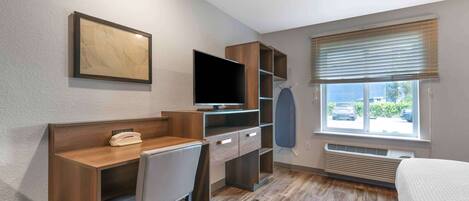 A hotel room with a wooden desk, chair, and television. There is a window with blinds and a view of the outside.