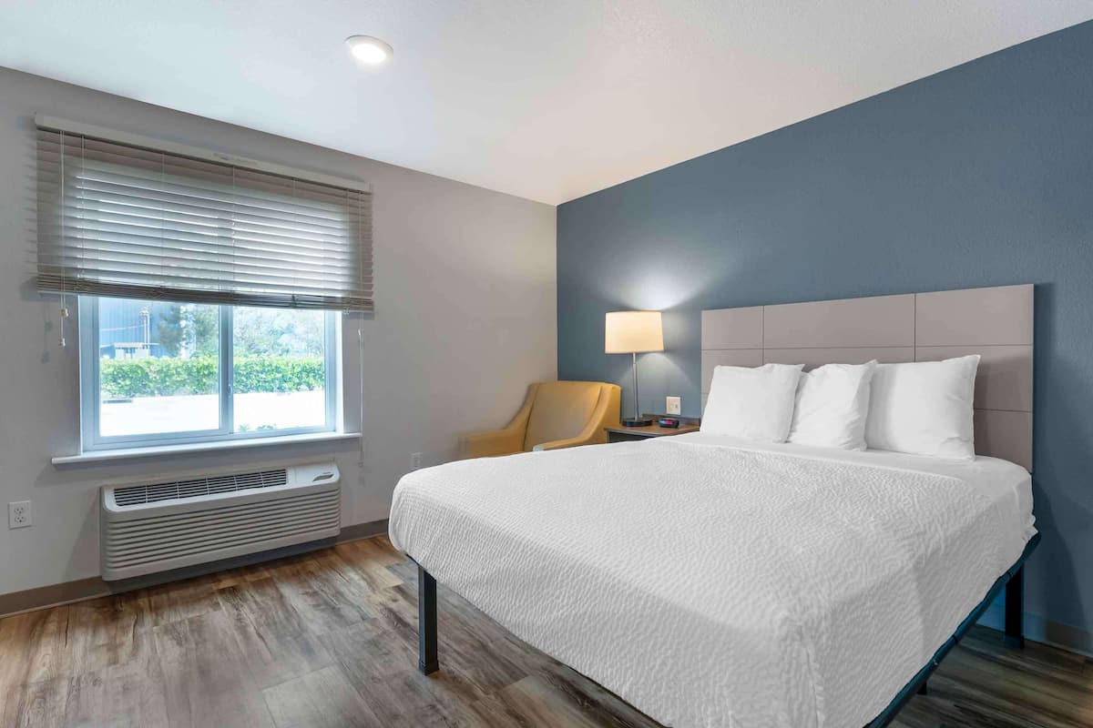 A modern hotel room with a large bed, a chair, a desk with a lamp, and a window with blinds.