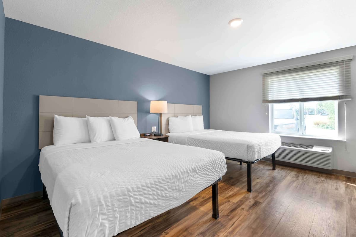 A hotel room with two beds, a nightstand with a lamp, a window with blinds, and a built-in heater.