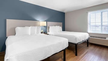 A hotel room with two beds, a large window with blinds, a wall-mounted heater, and a nightstand with a lamp.