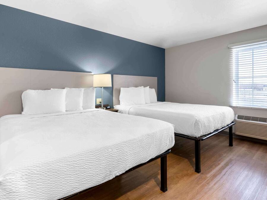 A hotel room with two beds, a large window with blinds, a wall-mounted heater, and a nightstand with a lamp.