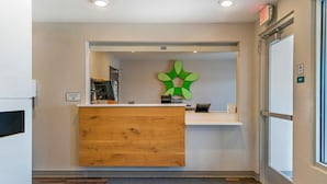 Una zona de recepción con un escritorio de madera, un adorno verde en la pared y una señal de salida.
