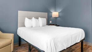 Una cama bien hecha con ropa de cama blanca y un cabecero, una lámpara de noche y una silla en una habitación con paredes azules y suelo de madera.