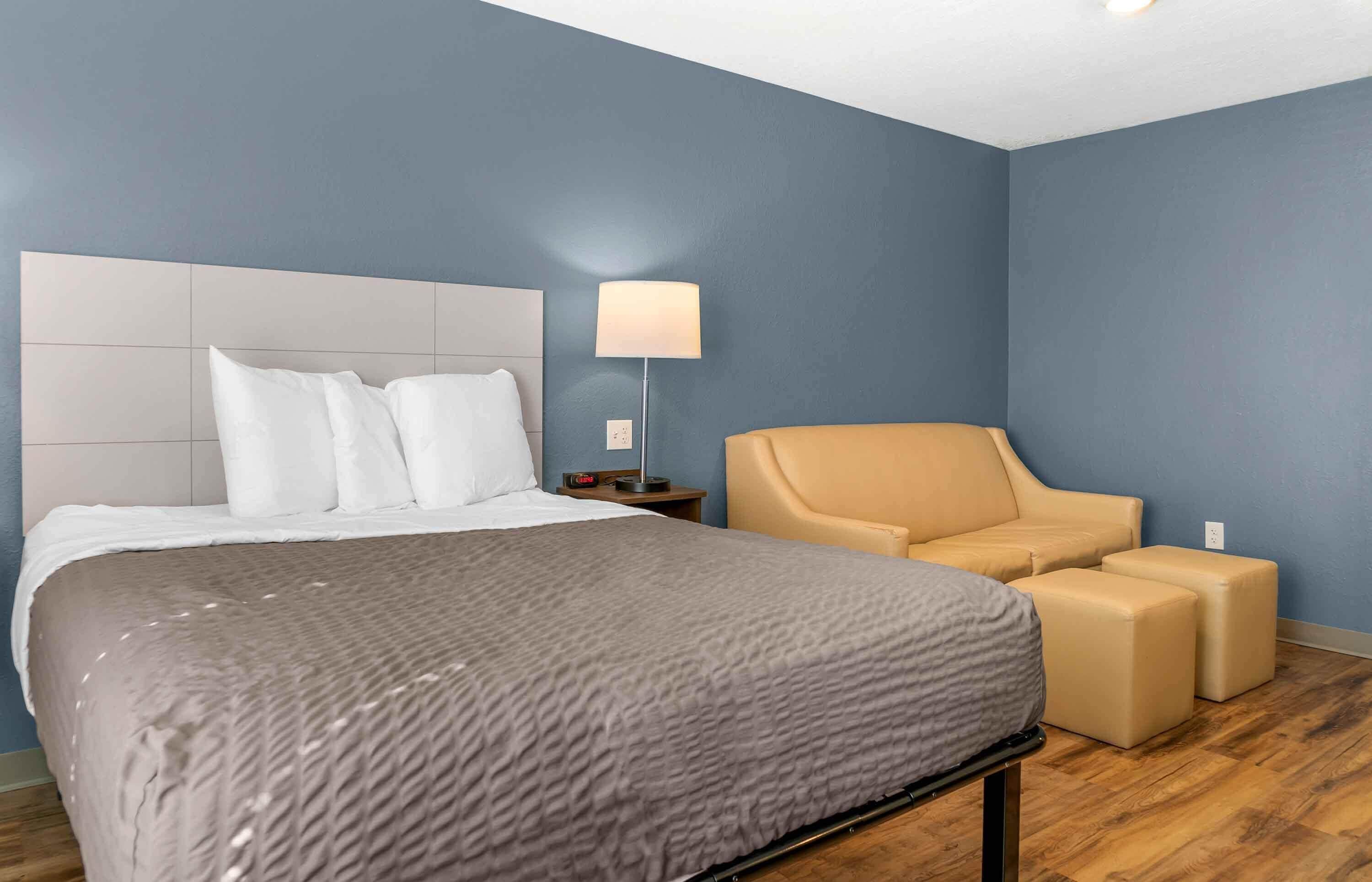 Una habitación de hotel con una cama, una mesita de noche, una lámpara y una zona de estar con un sillón y un reposapiés.