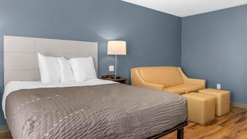 Una habitación de hotel con una cama, una mesita de noche, una lámpara y una zona de estar con un sillón y un reposapiés.