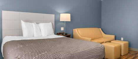 Una habitación de hotel con una cama, una mesita de noche, una lámpara y una zona de estar con un sillón y un reposapiés.