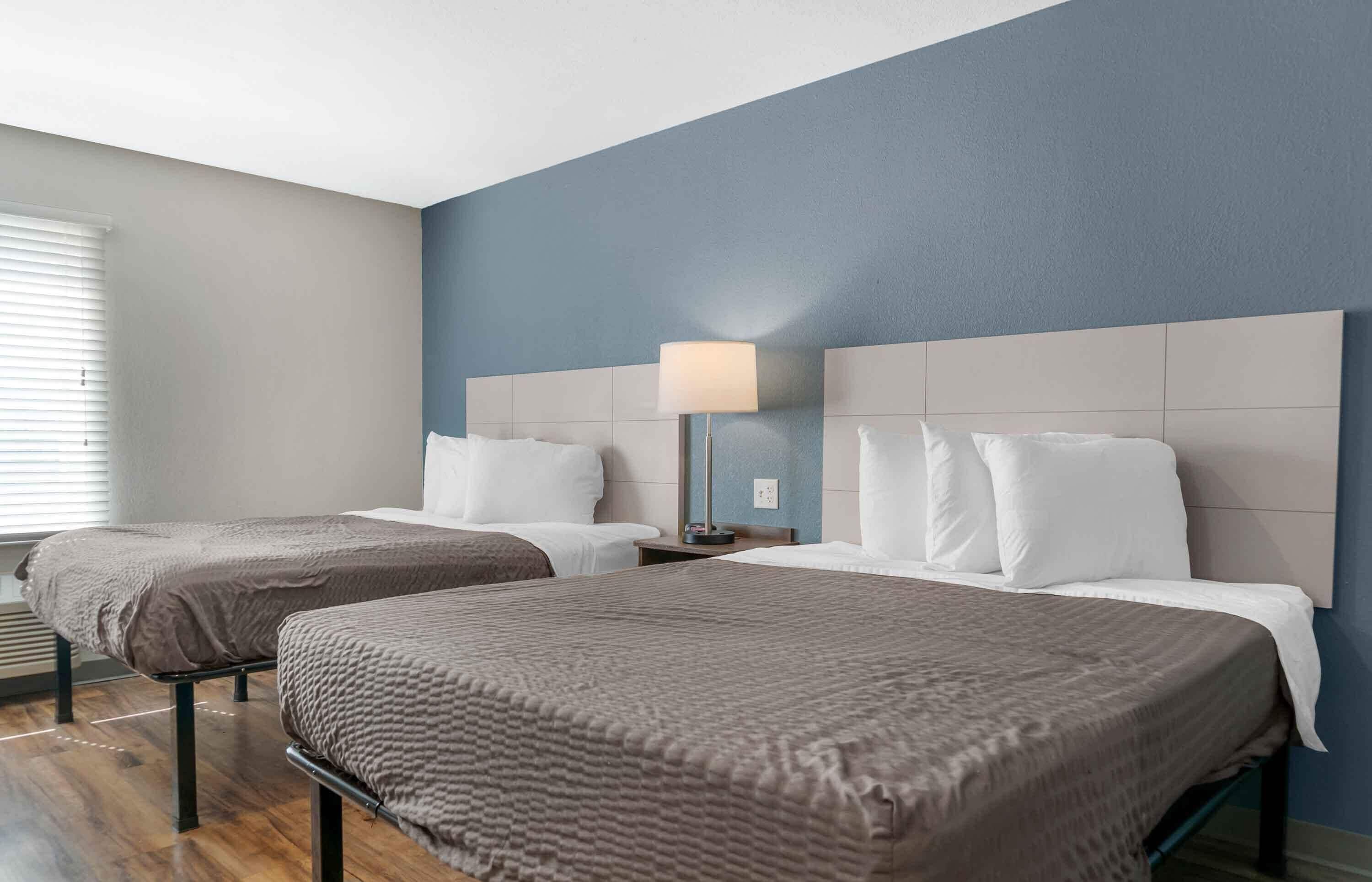 A hotel room with two beds, a nightstand, a lamp, and a window with blinds.