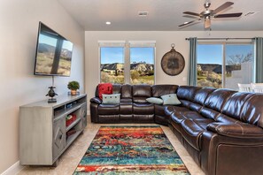 A living room with a sectional sofa, a flat-screen TV, a ceiling fan, and large windows with a mountain view. - Fairway Oasis (St. George)