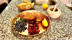 Een keuken met granieten aanrecht, roestvrijstalen apparatuur en een ontbijtarrangement met croissants, brood, blauwe bessen, druiven en een citroen.
