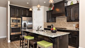 A kitchen with dark cabinetry, stainless steel appliances, a granite countertop, and two stools with green cushions. - The Painted Horse (St. George)