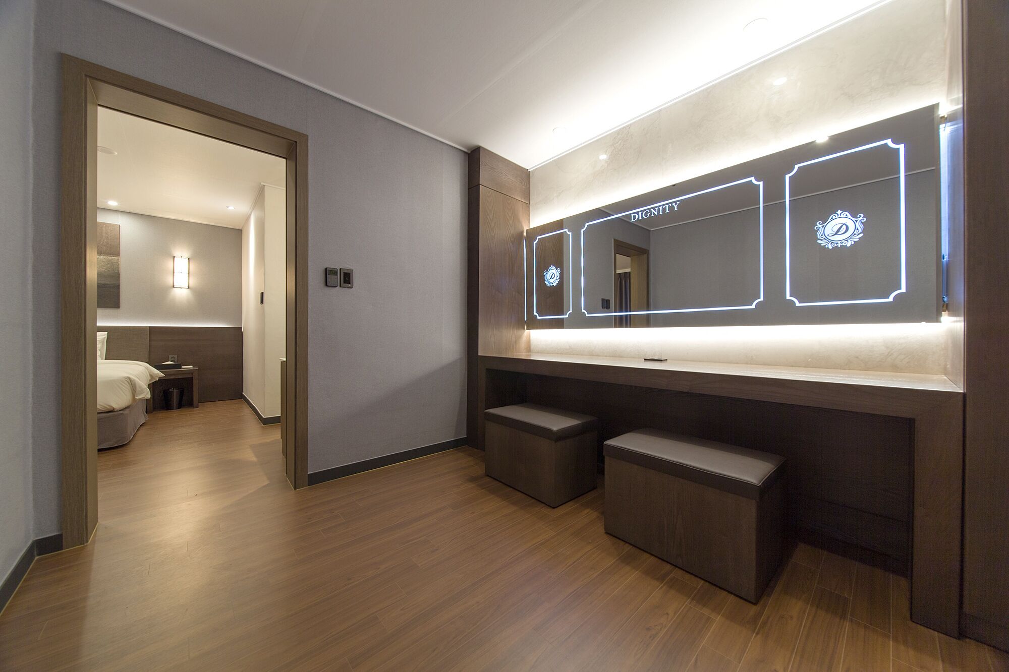 A modern hotel room with a wooden floor, a large mirror with illuminated edges, and a small bench.