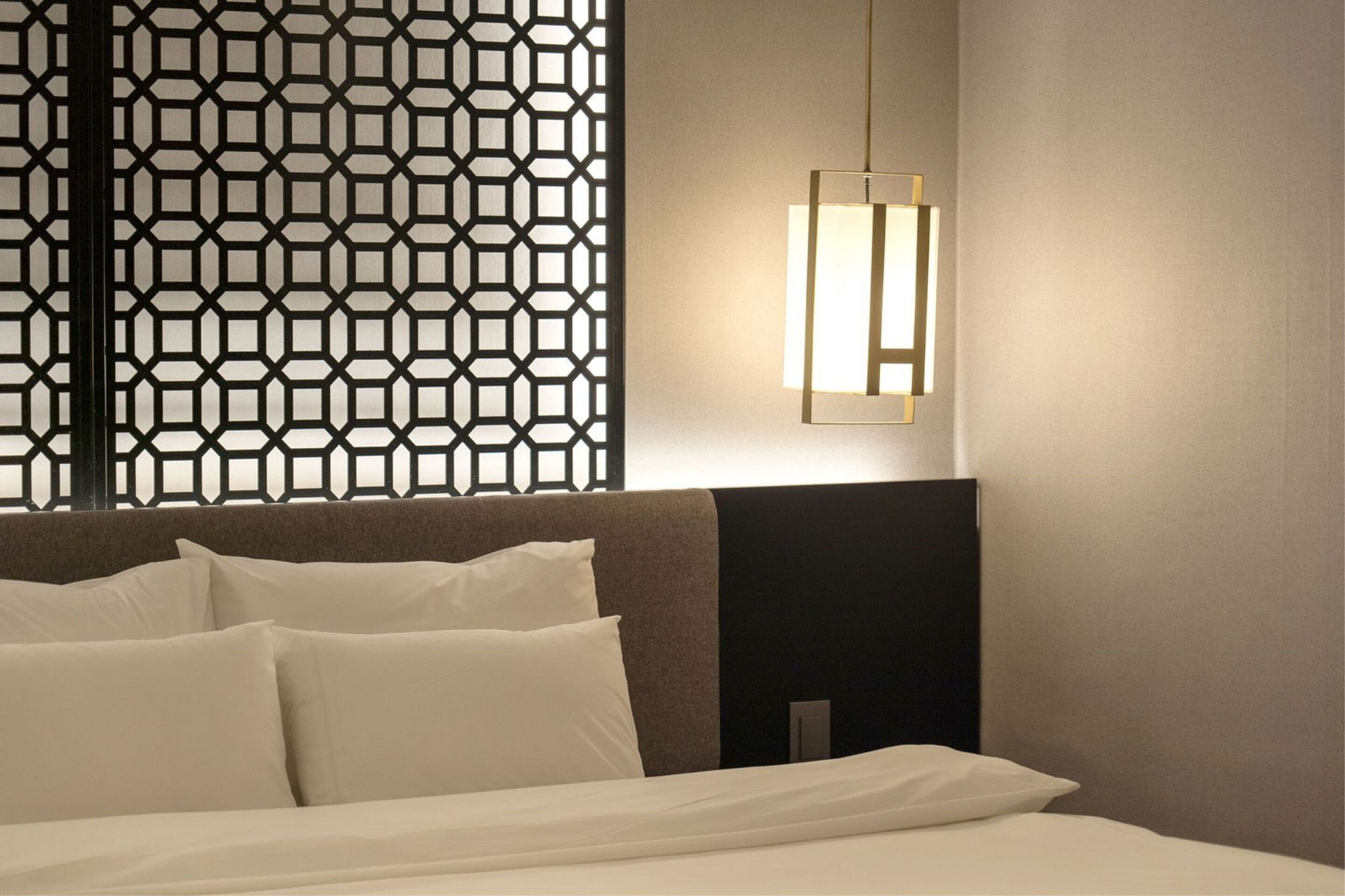 Habitación de hotel con cama, ropa de cama blanca, separador geométrico y lámpara fijada a la pared.