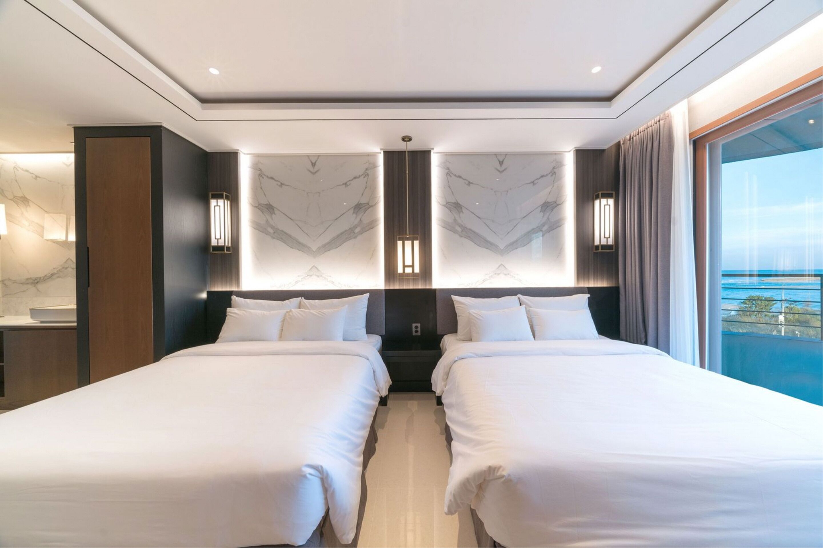 A modern hotel room with two beds, marble wall decor, and a large window offering a view of the sea.