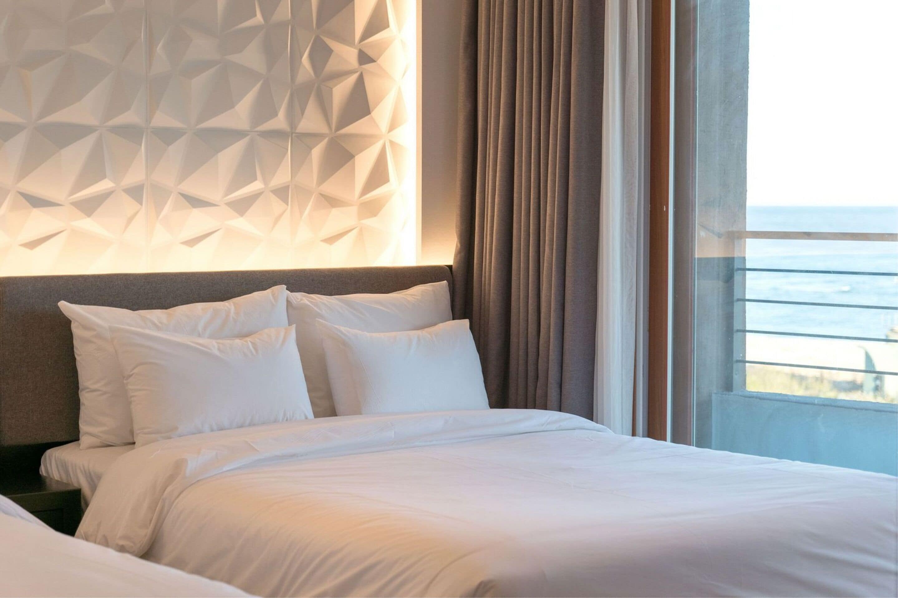 Quarto de hotel com cama, design geométrico na parede e vista para o oceano através de grandes janelas.