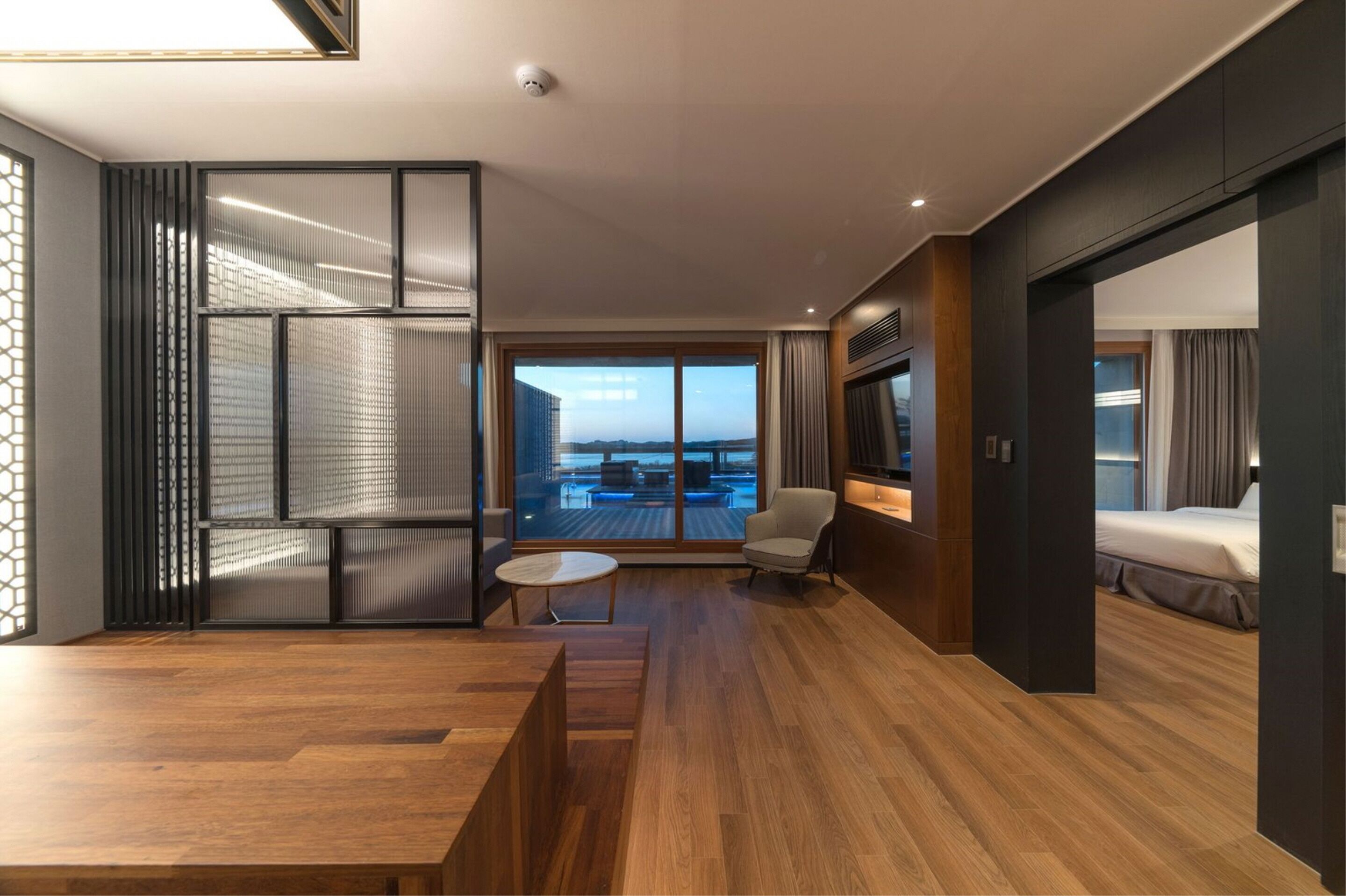 Una habitación de hotel moderna con suelo de madera, un ventanal con vistas a la ciudad, una zona de estar con una mesa redonda y una cama con cabecero oscuro.