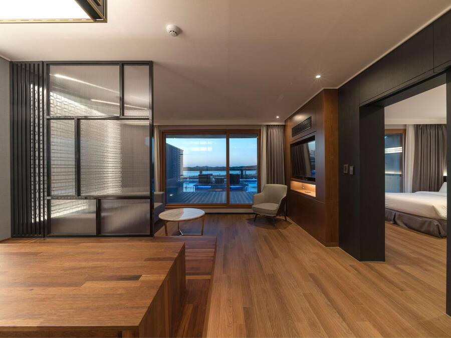 Una habitación de hotel moderna con suelo de madera, un ventanal con vistas a la ciudad, una zona de estar con una mesa redonda y una cama con cabecero oscuro.