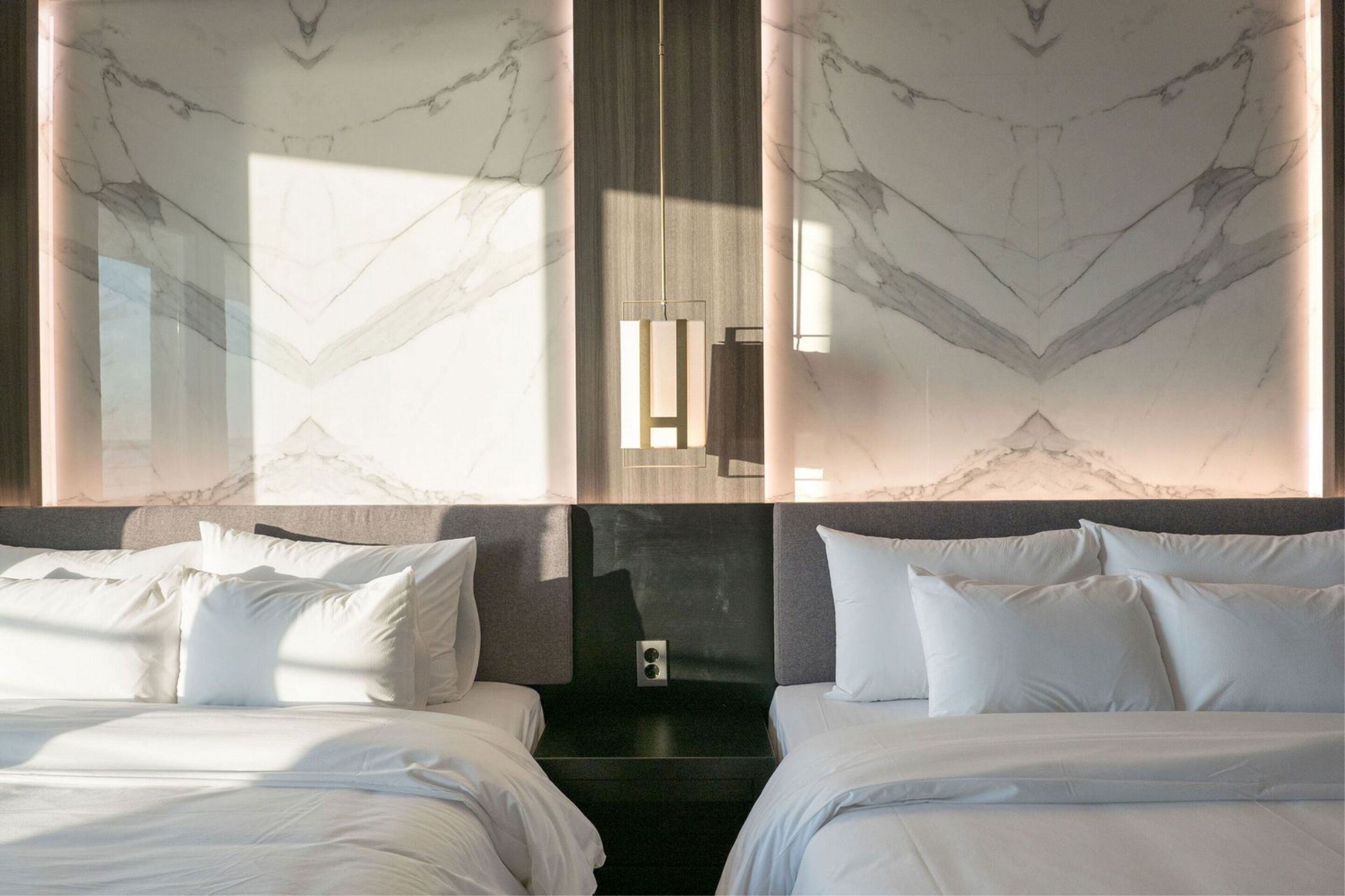 A hotel room with two beds, marble-patterned walls, a modern light fixture, and a view of the city.