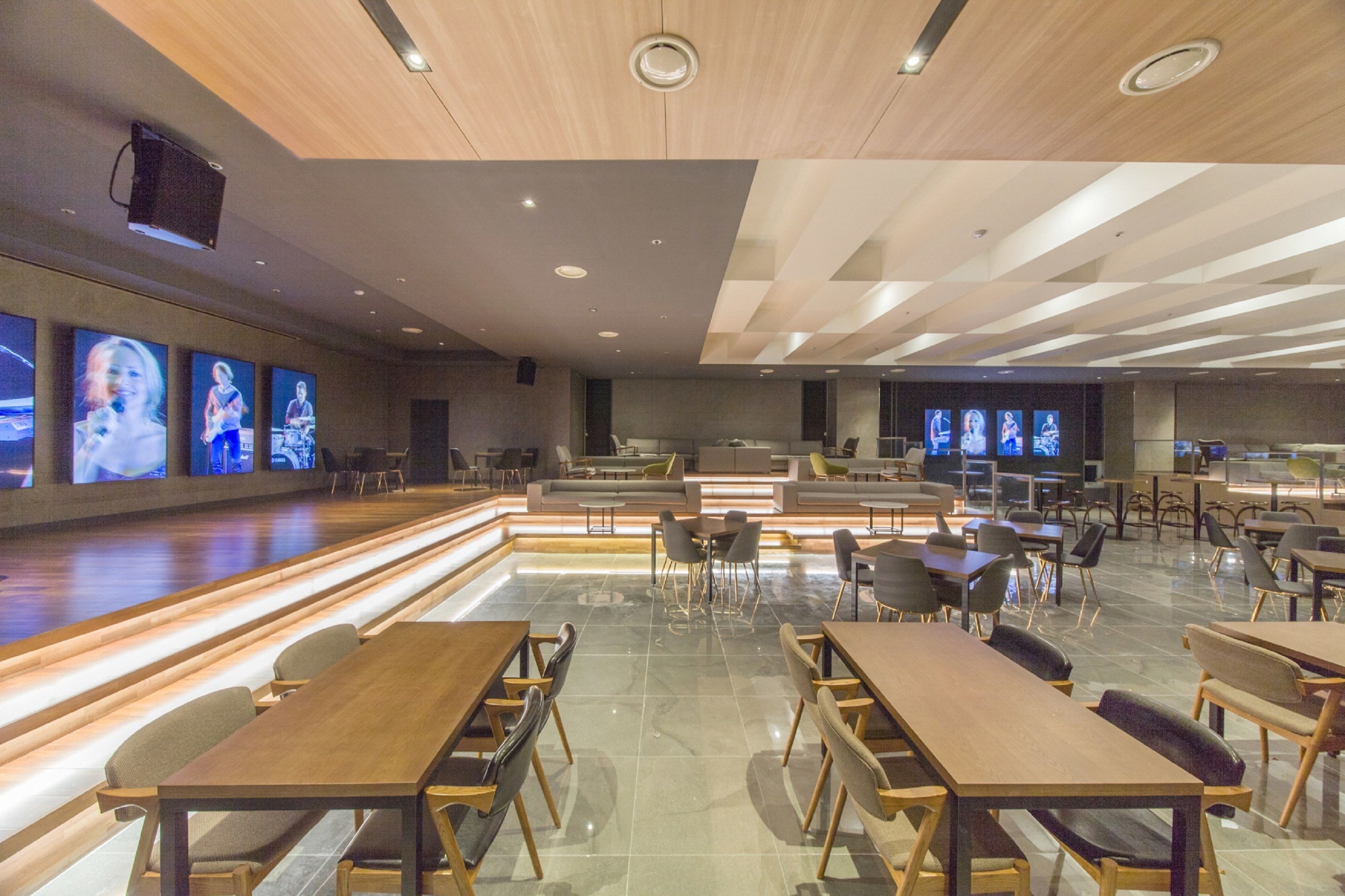 A spacious restaurant with multiple tables and chairs, a long counter, and a stage area with screens displaying images.