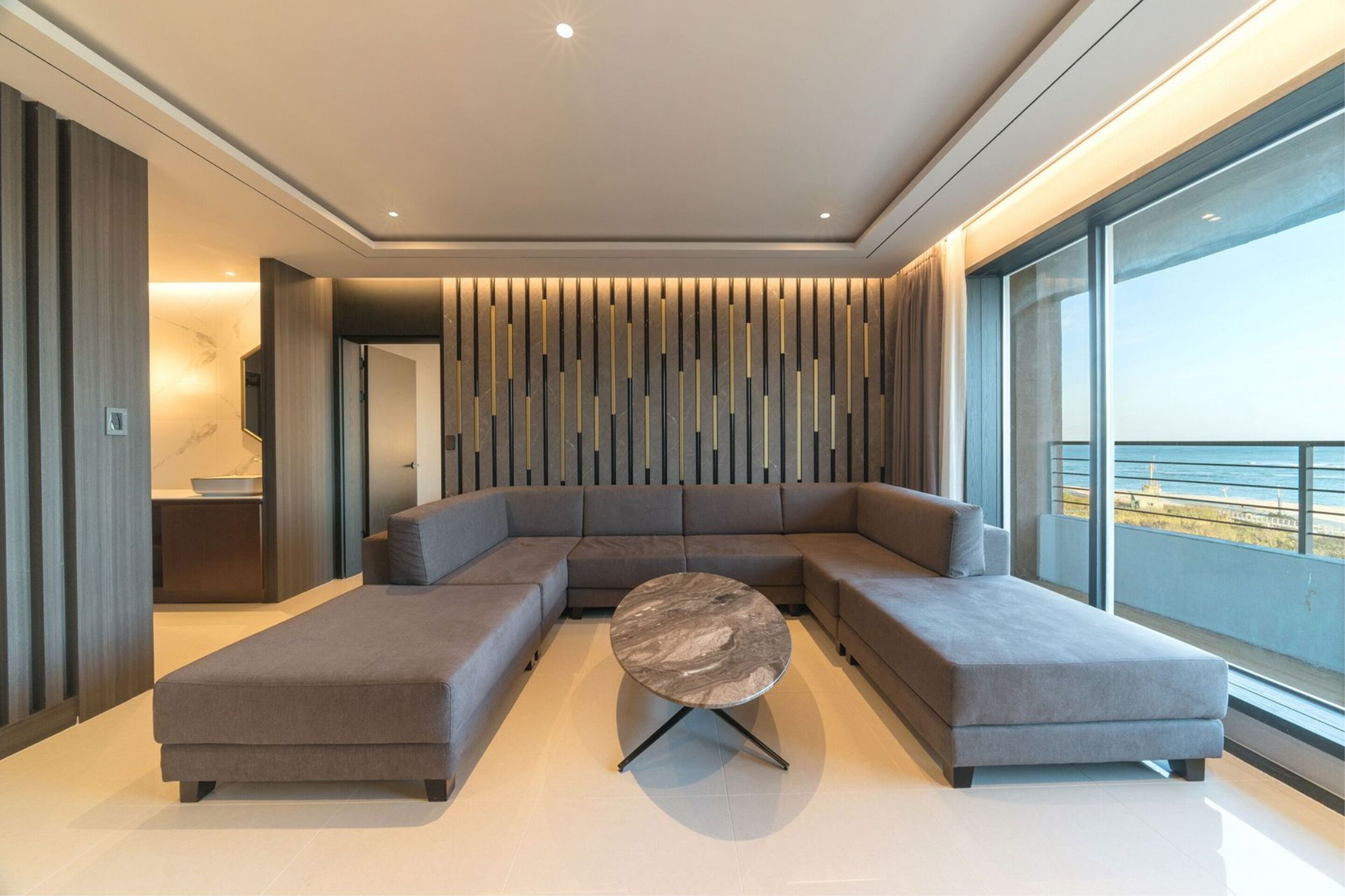 A modern living room with a sectional sofa, a round coffee table, and a large window offering a view of the sea.