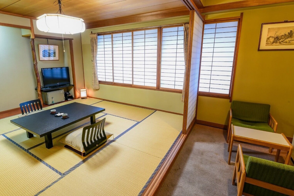 Ett traditionellt japanskt rum med tatamimattor på golvet, ett lågt bord, stolar, en tv och skjutdörrar.