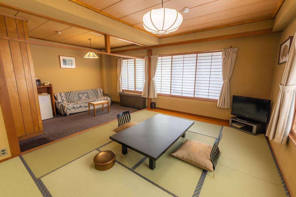 Ett traditionellt japanskt rum med tatamimattor på golvet, ett lågt bord, en soffa, en tv och en takfläkt.
