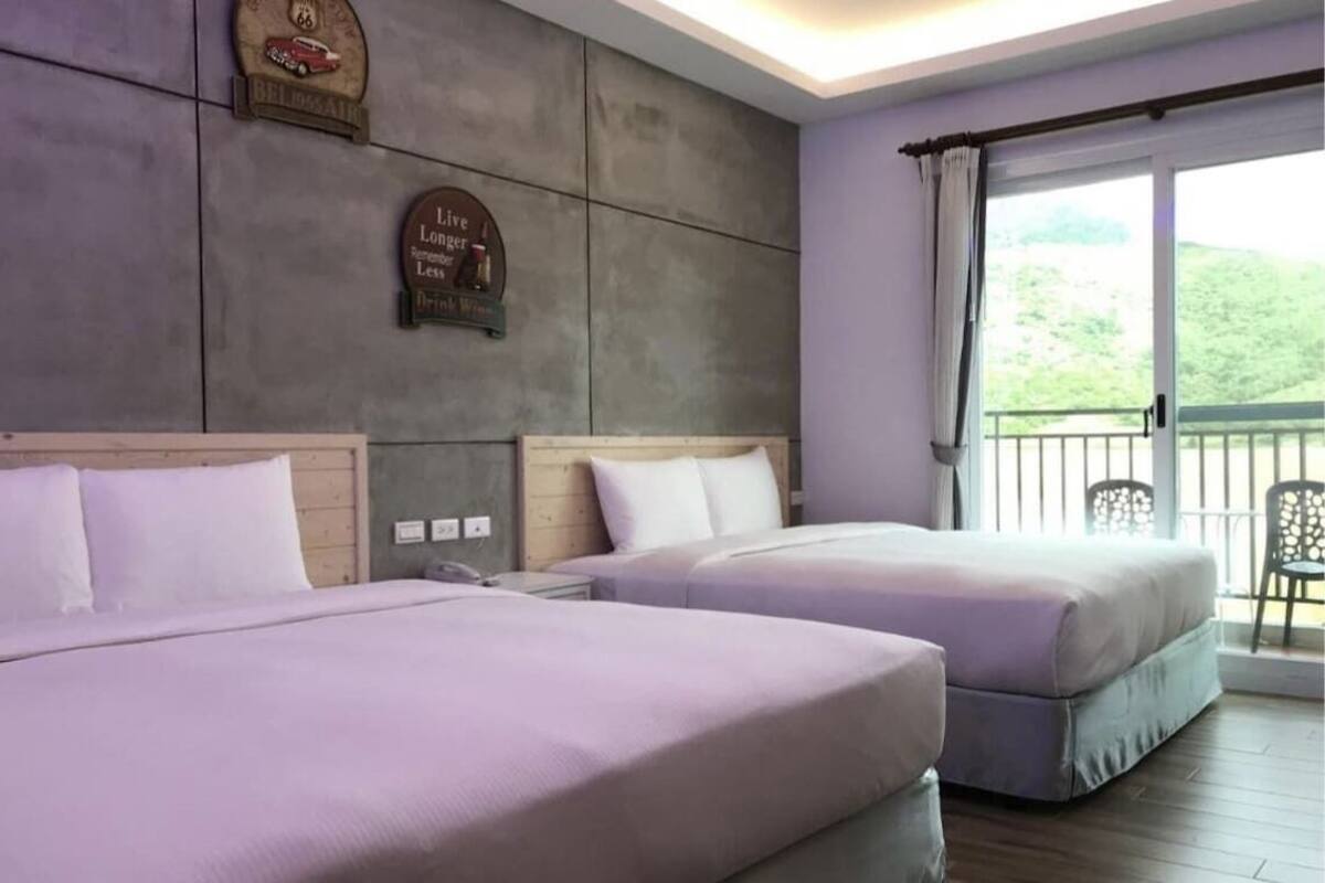 Habitación de hotel con dos camas, cabecera de madera, un reloj de pared y vistas a la vegetación desde un balcón.