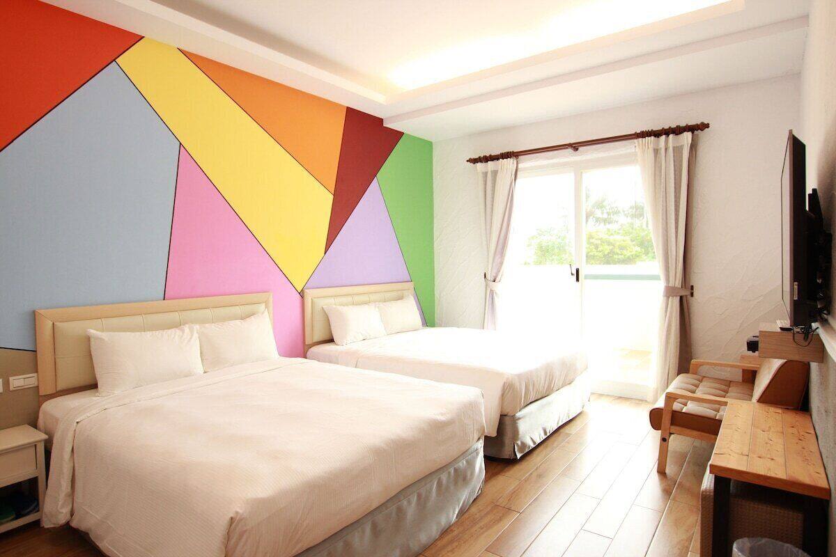 Habitación de hotel con dos camas, un escritorio de madera, un televisor y un llamativo diseño geométrico en la pared.
