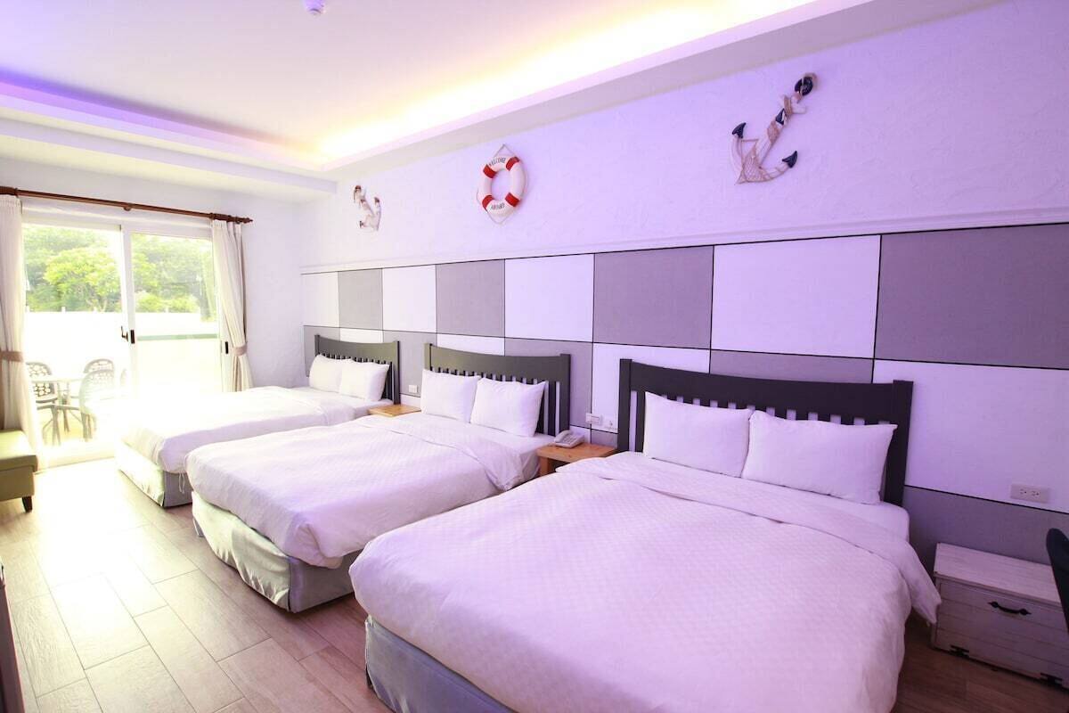 Habitación de hotel con tres camas, un ventanal grande y vistas a la vegetación.