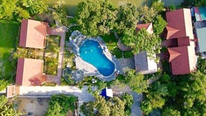 An aerial view of a property with a central swimming pool surrounded by lush greenery and multiple buildings with red roofs.