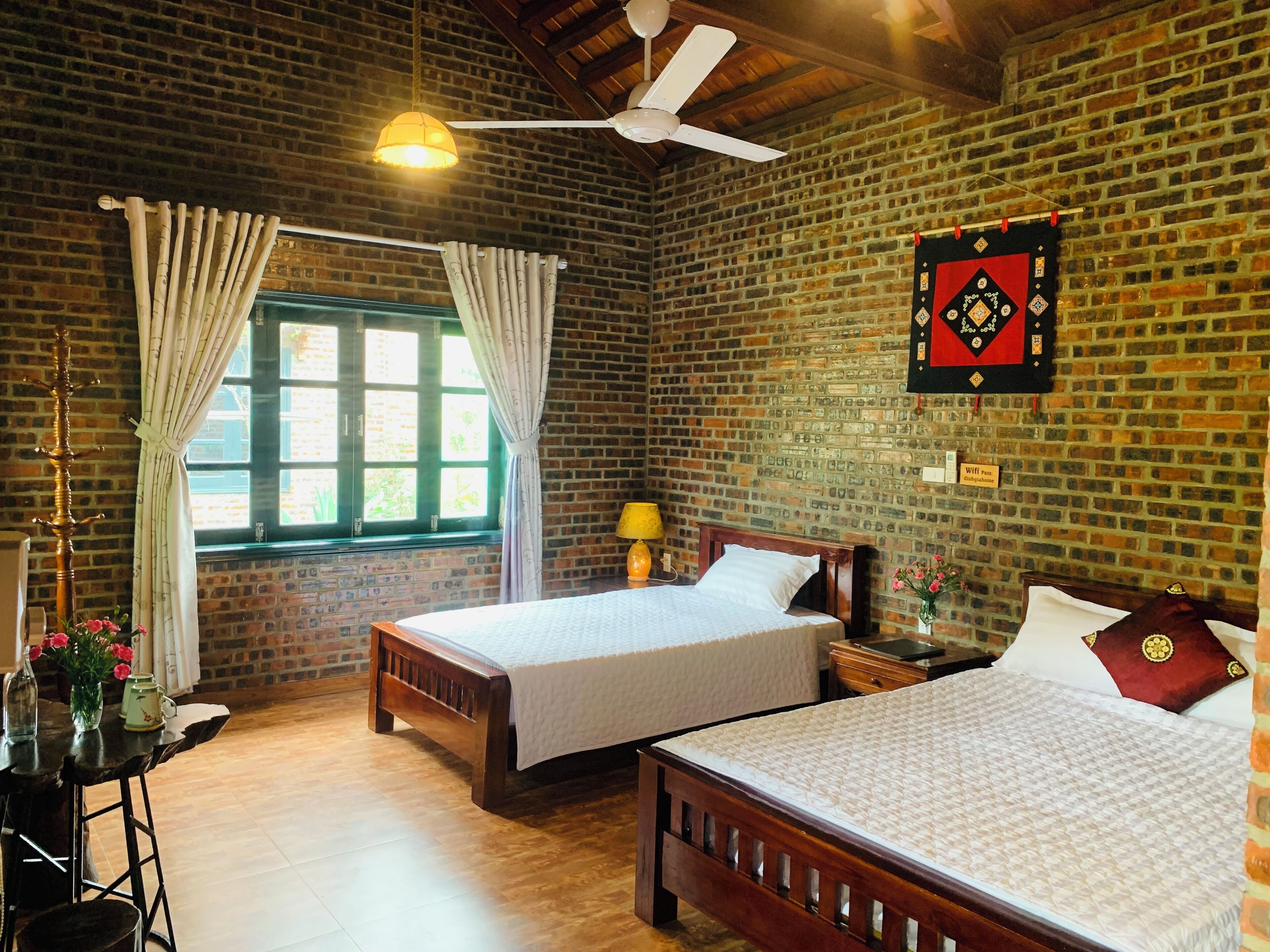 A room with two wooden beds, a brick wall, a window with curtains, a ceiling fan, and a sign indicating WiFi availability.