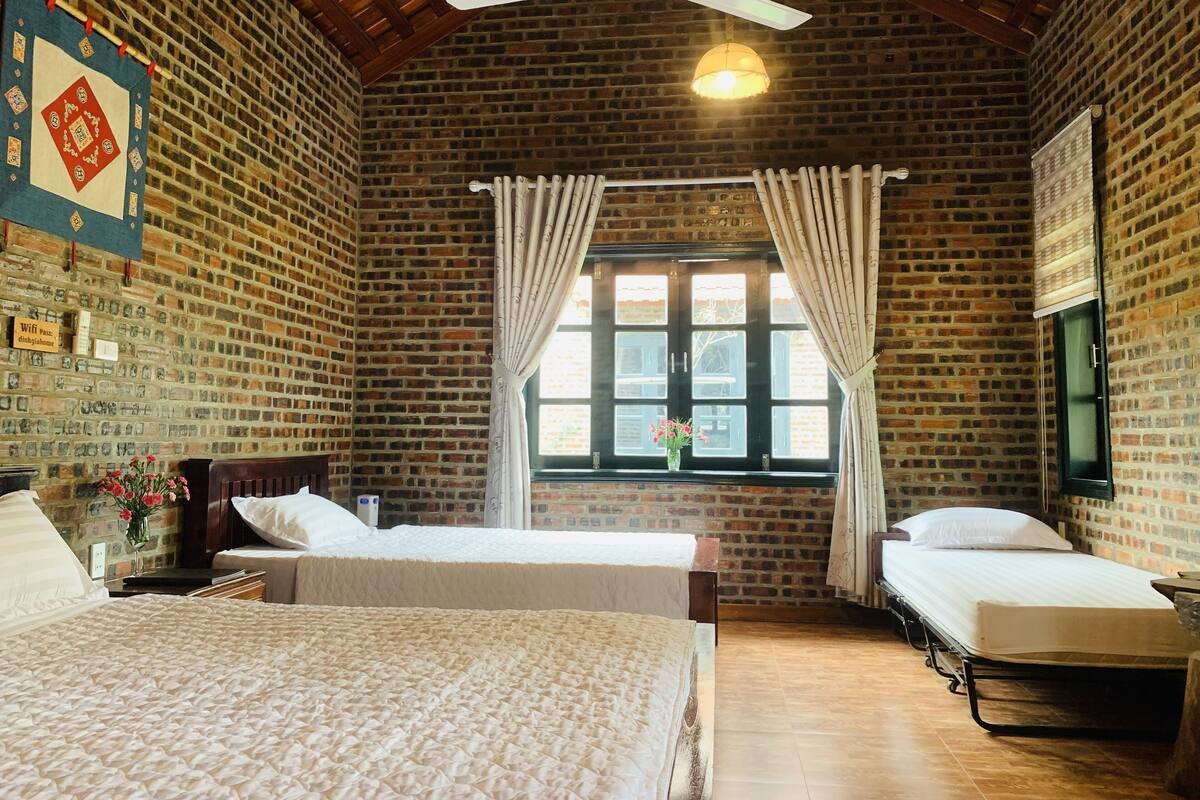 Una habitación con dos camas, una ventana con cortinas, un ventilador de techo y una pared de ladrillo.